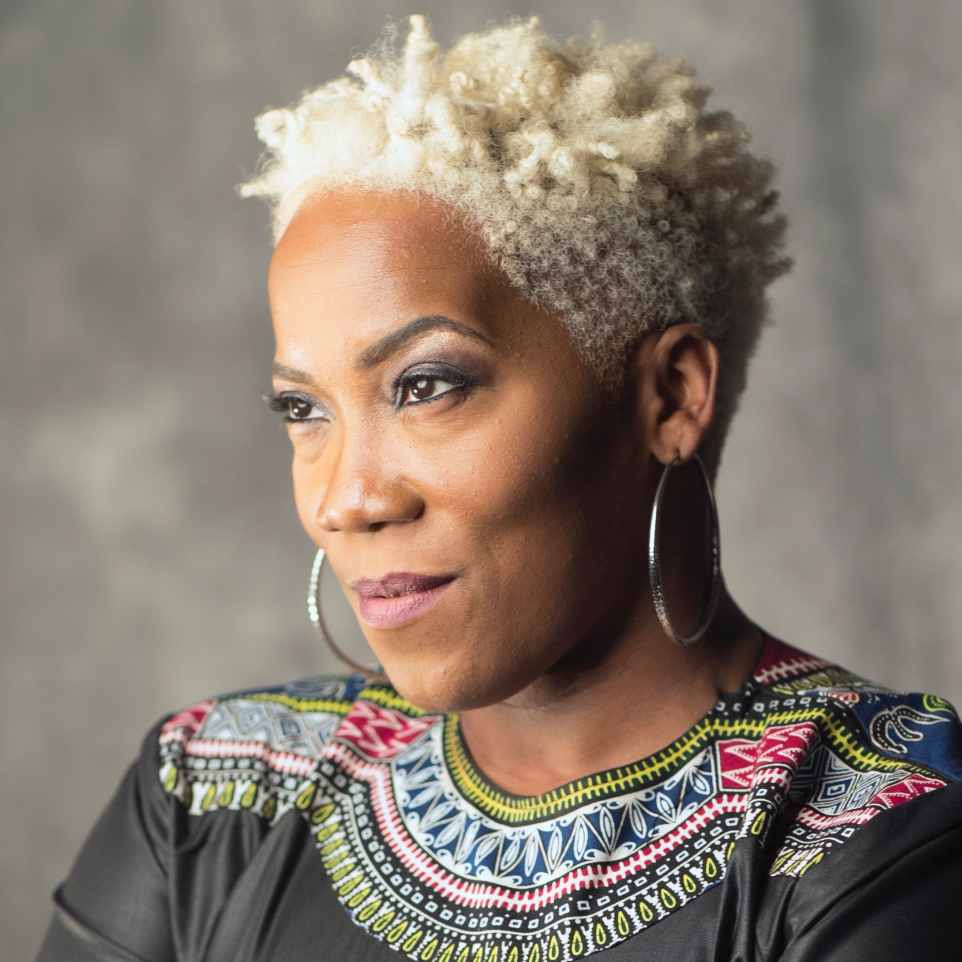 Portrait of JoVia Armstrong with short silver curls, hoop earrings, and a patterned top