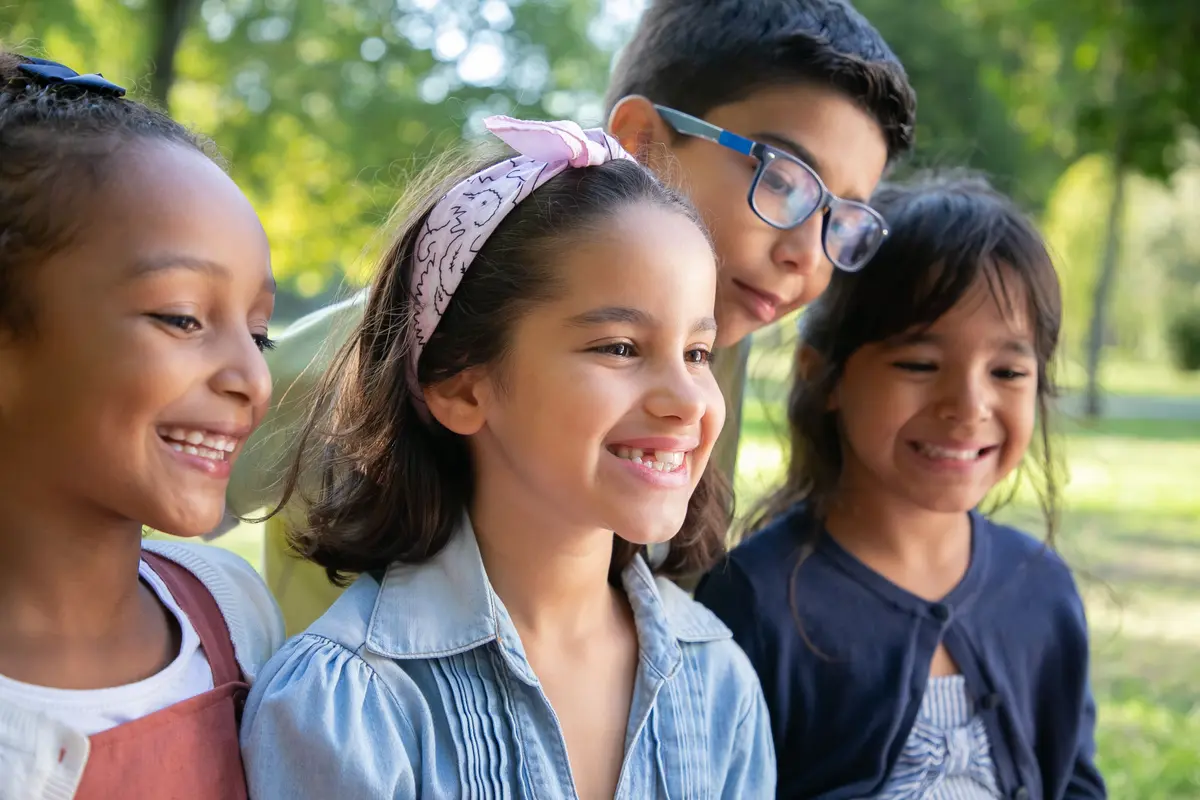 Four school-age children from diverse backgrounds smile together outdoors in a sunlit park.