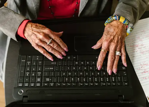 An older Black woman’s hands type on a laptop keyboard beside handwritten notes.