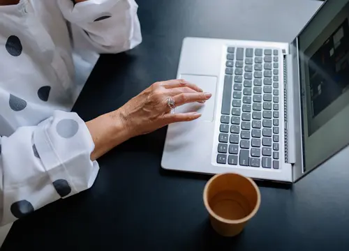 An older adult’s hand uses a laptop touchpad beside a cup of coffee.