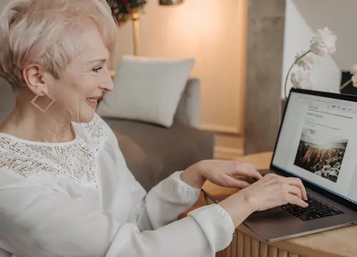 An older woman with short white hair smiles while using a laptop at home.