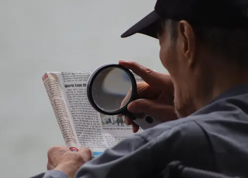 An older reader uses a magnifying glass to enlarge newspaper text.