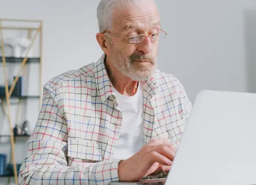 An older man wearing glasses works on a laptop at a bright desk.