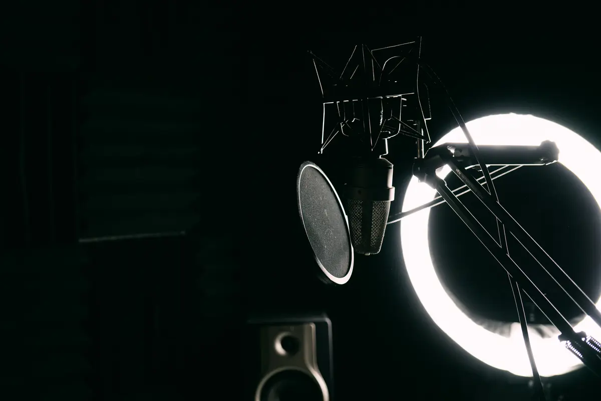 A studio condenser microphone and circular pop filter appear in silhouette before a bright white ring light in a dark recording space.