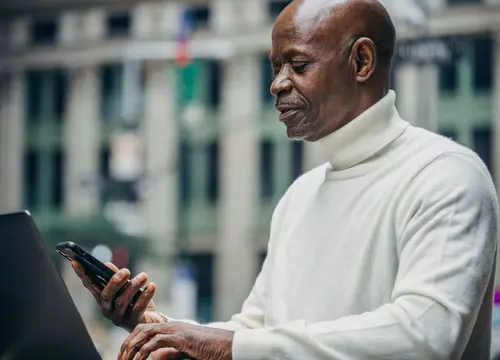 An older Black man uses a smartphone beside an open laptop in an urban setting.