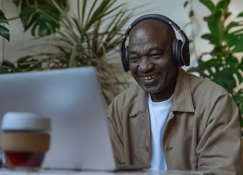 An older Black man wearing headphones smiles while using a laptop at home.