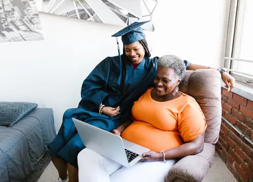 A younger Black woman helps an older Black woman use a laptop while they sit together.
