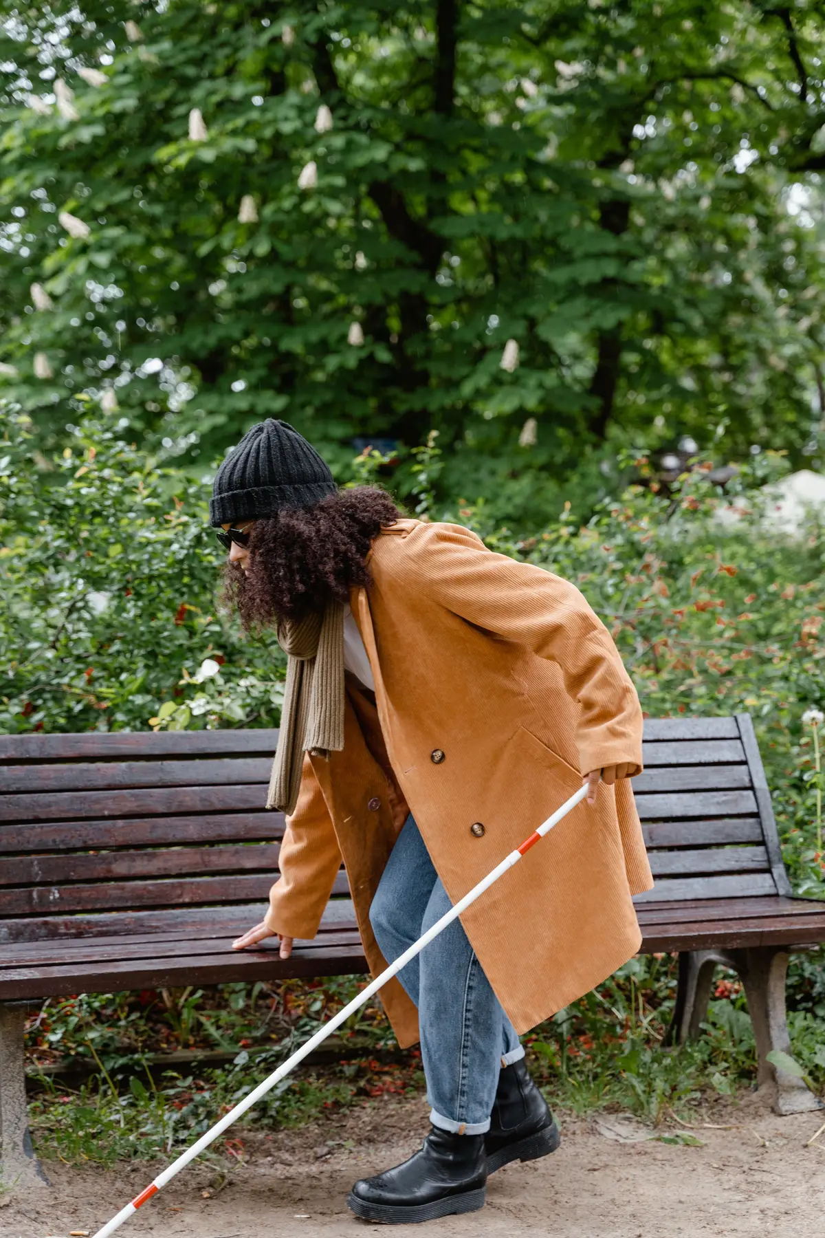 A woman wearing sunglasses and a tan coat uses a white cane as she approaches a wooden bench on a tree-lined path.